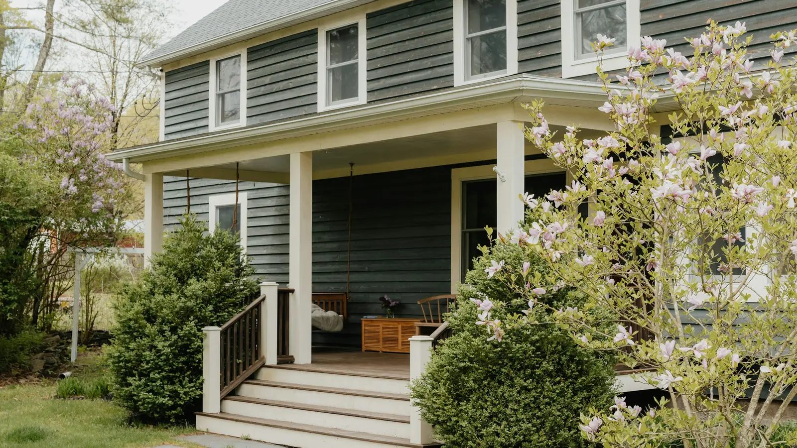 Covered front porch of a gray two-story house with steps and potted plants