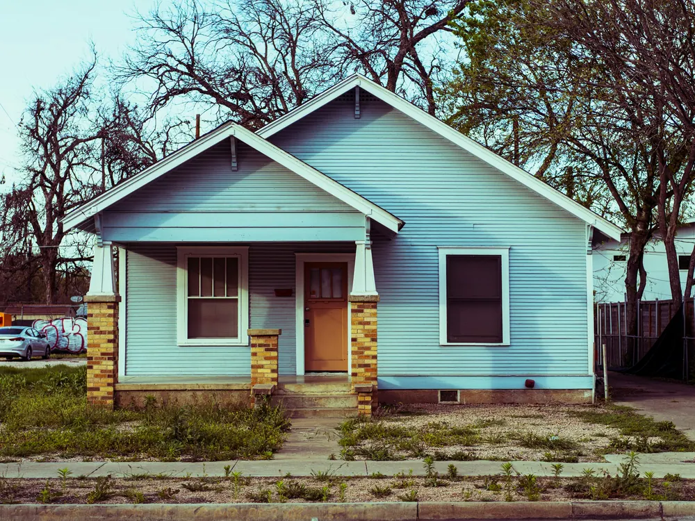 Small light-blue bungalow with stone porch columns and a worn front yard