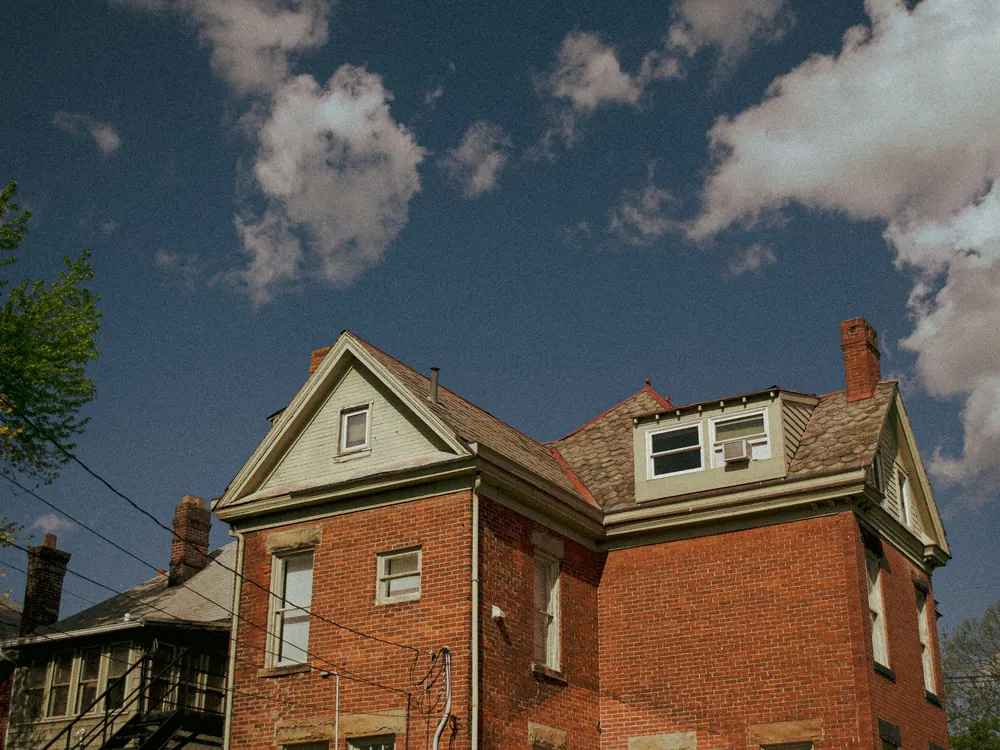 Older red-brick two-story house with dormer windows under a cloudy sky