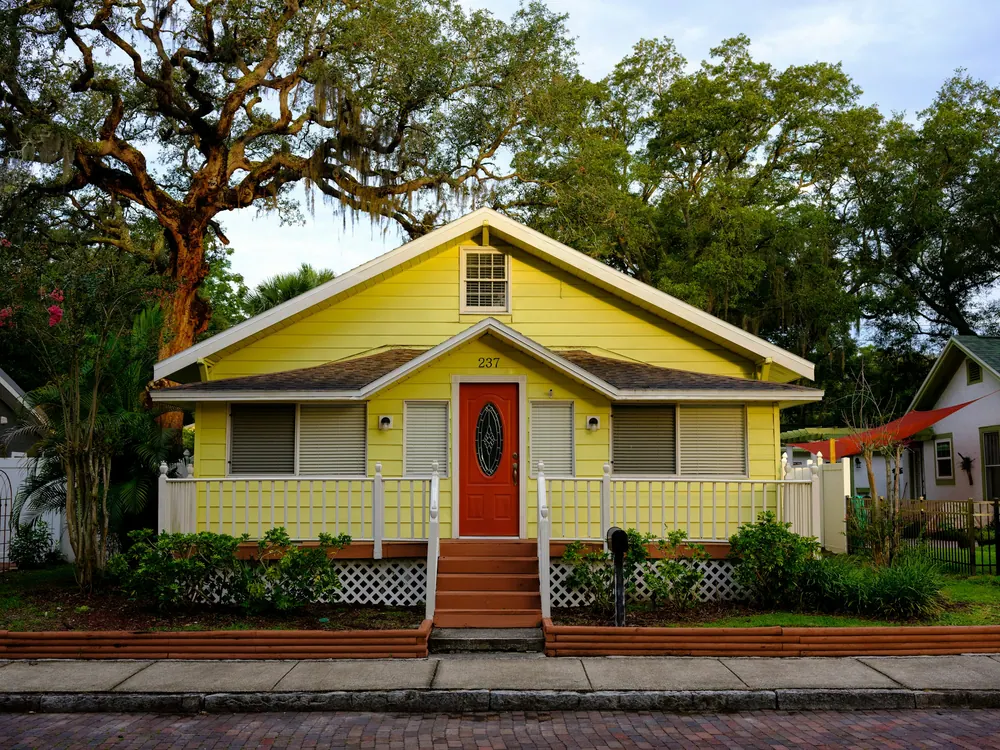 Yellow craftsman bungalow with a red door under large shade trees