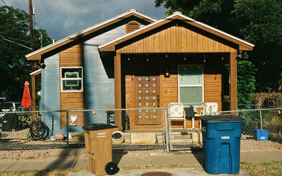 Small rental house behind a chain-link fence with trash bins and clutter in the yard