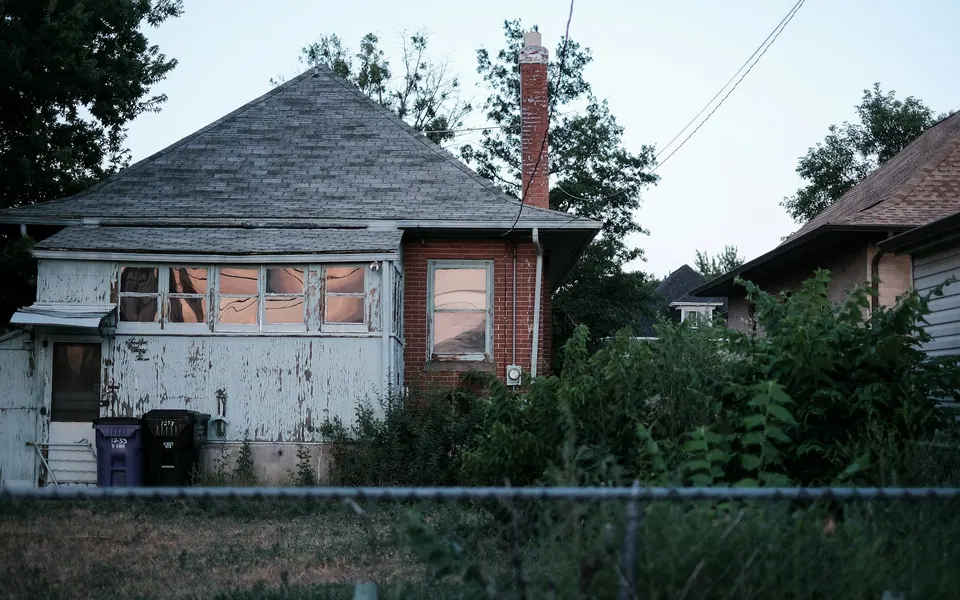 Worn house with a broken window, aging roof, and overgrown shrubs — deferred maintenance